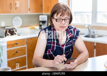 Horizontale Nahaufnahme einer kaukasischen Frau, die in ihrer Küche mit ihrem Handy sitzt. Stockfoto