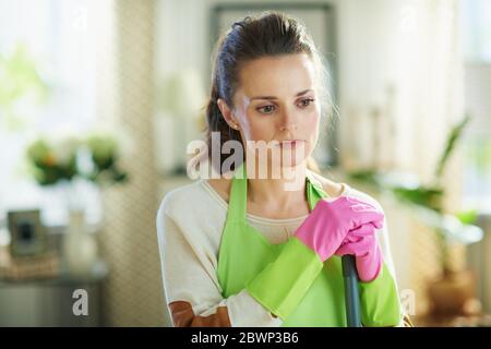 Nachdenkliche junge Frau in grüner Schürze und rosa Gummihandschuhe mit Mopp in dem modernen Haus an sonnigen Tag. Stockfoto