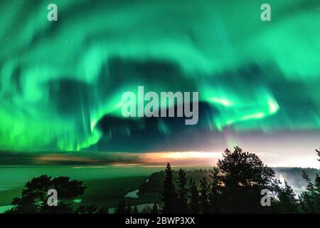 Blick auf die leuchtend grüne Aurora, die über die schwedische Nebelwaldlandschaft in den Bergen glänzt, Lichtstrahlen aus einem Dorf und Nordlichter färben Himmel in di Stockfoto