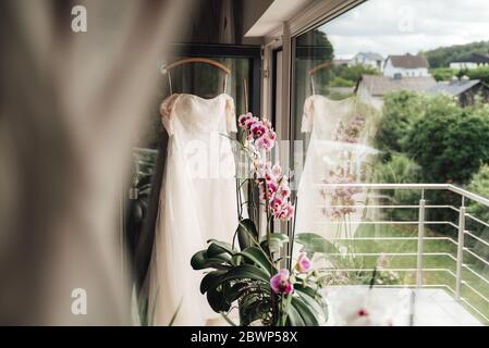 Weißes Hochzeitskleid auf Kleiderbügel an Balkontür gehängt. Halbreflektiertes Kleid in großem Fenster. Bunte blühende Orchideenblume. Stockfoto