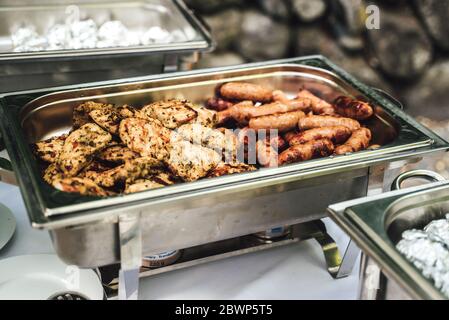 Edelstahlpfanne mit gewürzten Hühnchen-Steaks und gegrillten Würstchen. Selbstbedienungs-Buffet. Feier, Party, Geburtstag oder Hochzeit Konzept. Stockfoto