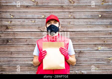 Versand Mann in roter Uniform Schutzmaske tragen und Handschuhe hält eine Papiertasche auf Holzhintergrund. Covid-19 Pandemie sicherer Lieferservice. Stockfoto