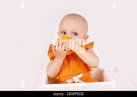 Glückliches Baby Mädchen, das im Hochstuhl sitzt und Orangen in einer weißen Küche isst. Gesunde Ernährung für Kinder. Bio-Früchte als erste feste Nahrung für Säuglinge Stockfoto