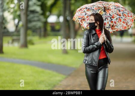 Ein maskiertes Mädchen läuft auf der Straße. Ein Mädchen in einer Schutzmaske geht im Park mit einem Regenschirm im Regen. Coronavirus-Infektion COVID-19 Stockfoto