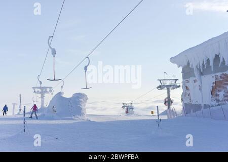 Zwei Skifahrer gehen eine Piste hinunter, nachdem sie von Skiliften aus gelandet sind, die sich über den Wolken in den hellblauen Himmel erheben. Sehr kalte und schneereiche Bedingungen. Stockfoto