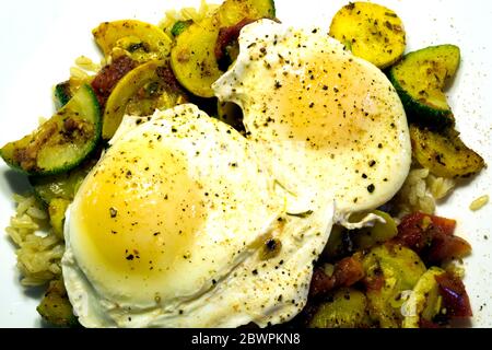 Zwei pochierte Eier liegen auf einem Bett aus gebratenen Zucchini, Sommer-Kürbis, Tomaten und braunem Reis. Stockfoto