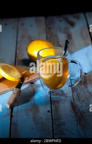 Appetitliche Tasse Tee mit einer Zitronenscheibe umgeben von einem Schneidebrett mit frisch geschnittener Zitrone und einem Messer und einer ganzen Zitrone auf einem Holztisch. Entspannen Stockfoto