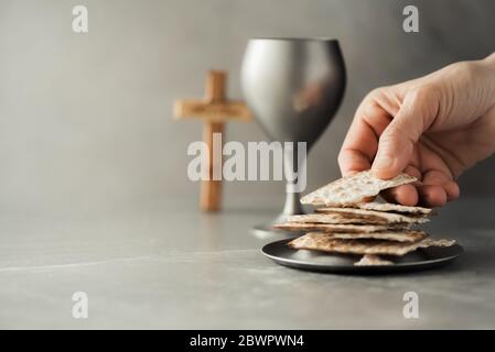 Hände mit Kelch und Kommunion Matzo Brot, Holzkreuz auf grauem Hintergrund. Christliche Kommunion zur Erinnerung an das Opfer Jesu. Osterpassah Stockfoto