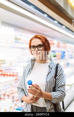 Junge Frau in Brillen und gestreiftem Hemd, die eine Flasche Milch in den Händen hält, während sie in einem modernen Supermarkt nachdenklich in die Kamera schaut Stockfoto