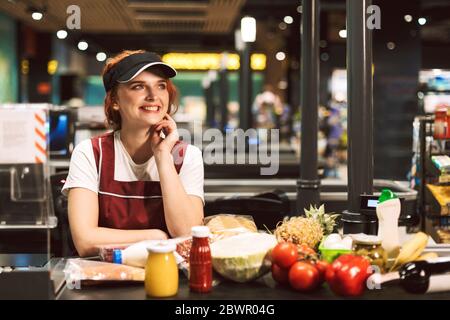 Junge fröhliche weibliche Kassiererin in Uniform freudig Blick beiseite mit Produkten auf der Kasse in der Nähe während der Arbeit in modernen Supermarkt Stockfoto