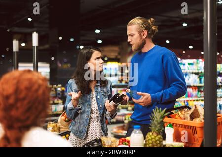 Ein junges Paar in der Nähe der Kasse, das emotional etwas im Supermarkt diskutiert Stockfoto