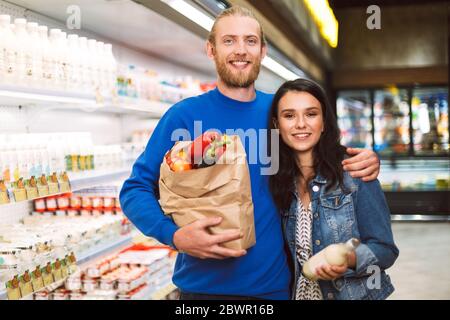 Junge schöne Paar mit Papier Lebensmitteltasche voller Produkte glücklich in der Kamera in der Milchabteilung des Supermarktes suchen Stockfoto