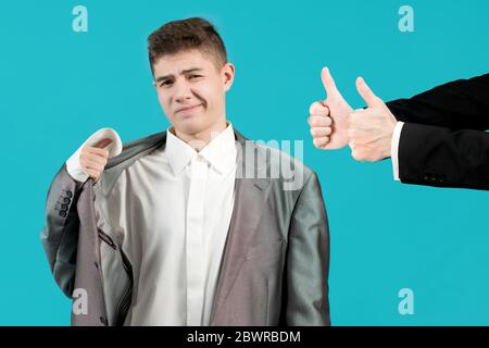 Der Teenager fühlt sich in der Kleidung eines erwachsenen Mannes unwohl, aber die Hände auf der Seite genehmigen mit einer Geste Daumen nach oben Stockfoto