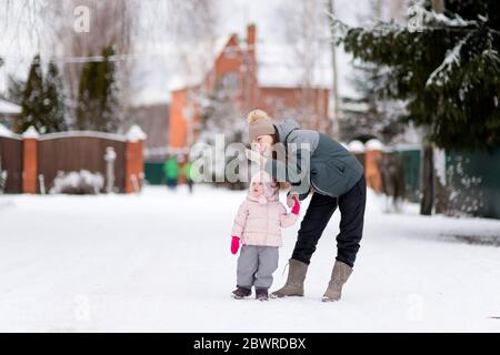 Eine junge Mutter zeigt auf etwas zu ihrer kleinen Tochter, mit der sie an einem Wintermorgen durch ein verschneiten Dorf geht Stockfoto