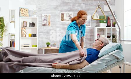 Krankenschwester hilft einem alten kranken Mann, der im Krankenhausbett in einem Pflegeheim liegt Stockfoto