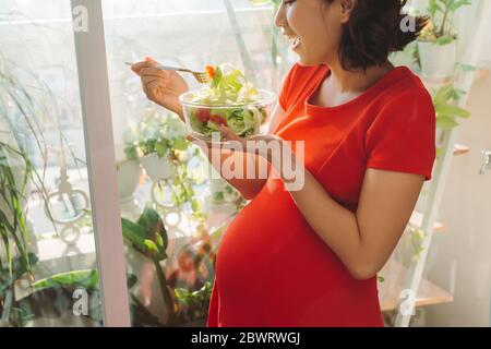 Voller Vitamine. Energisch gut aussehende schwangere Frau, die ihr Fleisch isst, während sie Teller in der Hand trägt und sich am Fenster entspannt Stockfoto
