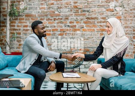 Zwei selbstbewusste Geschäftsleute, Afrikaner und Muslimin in weißem Hijab, die sich bei einem Treffen im Büro die Hände schütteln, nachdem sie erfolgreich umgebracht wurden Stockfoto