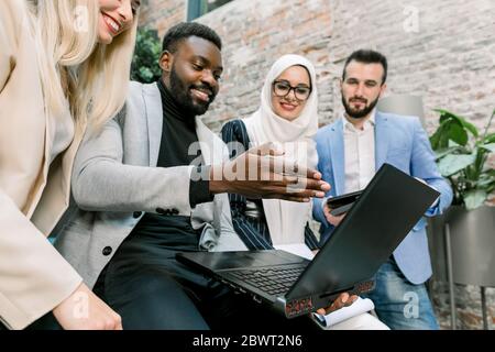 Team von multiethnischen verschiedenen Geschäftsleute, zwei Männer und zwei Frauen, stehen in modernen Büro, diskutieren und arbeiten zusammen, mit Laptop und Stockfoto