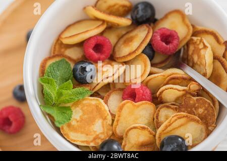 Frühstück weiße Schüssel mit winzigen Pfannkuchen Müsli mit Heidelbeeren, Himbeeren und Minze auf Holzhintergrund, Nahaufnahme, selektive Fokus Stockfoto