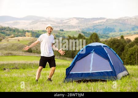 Junger Mann mit Hands Up Camping Zelt mit Blick auf die atemberaubende Aussicht auf den Berg mit blauem Himmel. Stockfoto