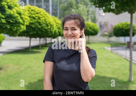 Indisches Mädchen hört Musik in kabellosen Kopfhörern, auf einem Hintergrund des grünen Parks. Junge Sportlerin in kabellosen Kopfhörern, wirbelt, streuende Haare Stockfoto