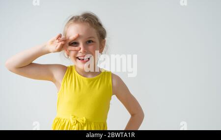 Freudige kleine Mädchen mit glänzenden entzückenden Lächeln suchen durch ihre zwei gespreizten Finger Hand auf Hüfte. Freigestellte Aufnahme isoliert auf weißem Kopierbereich Stockfoto