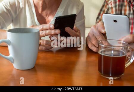 Ältere Paare, die zu Hause moderne Mobiltelefone benutzen und dabei Tassen heiße Getränke genießen. Konzept der älteren Menschen und Technik, persönliche Entwicklung... Stockfoto