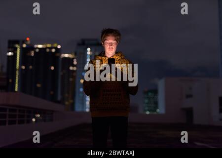 Junger schöner Mann mit Telefon gegen Blick auf die Stadt bei stürmischem Wetter in der Nacht Stockfoto
