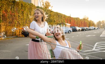 Porträt von zwei lachenden Mädchen Blasen Seifenblasen während der Fahrt in Shopping-Trolley auf dem Parkplatz in der großen Mall Stockfoto