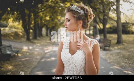 Getöntes Porträt von eleganten blonden Braut in weißem Kleid posiert auf langen Gasse im Park Stockfoto