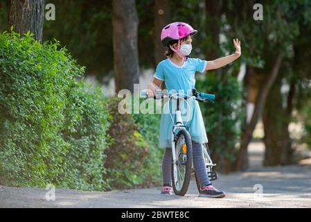 Nettes kleines Mädchen mit Maske auf Gesicht befreien ein Fahrrad und grüßen andere Kinder aus der Ferne. Stockfoto