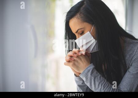 Porträt einer Frau mit chirurgischer Maske, die neben dem Fenster betet Stockfoto
