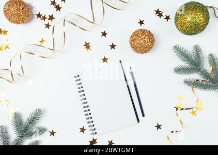 Draufsicht leeres Notizbuch unter goldenen Weihnachtsbaumkugeln, sternförmigen Konfetti, Bändern und grünen Fichtenzweigen auf weißem Hintergrund, flaches Lay. Stockfoto