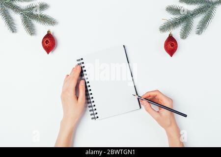 Weibliche Hände schreiben in leerem Notizbuch in der Nähe von Zweigen von Weihnachtsbäumen mit roten Ornamenten auf weißem Tisch, Draufsicht. Stockfoto