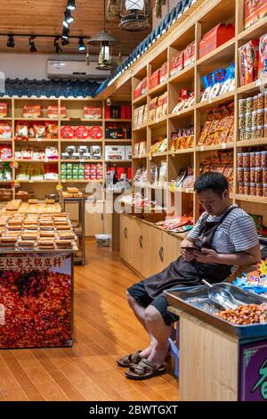 Chongqing, China - August 2019 : gelangweilter chinesischer Verkäufer überprüft sein Smartphone während er sitzt und auf Kunden in seinem Snack-und Lebensmittelgeschäft wartet Stockfoto