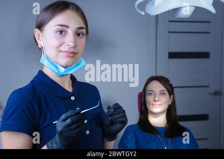 Porträt eines jungen, schönen Zahnarztarztes in Maske, lächelnd, hält zahnmedizinische Instrumente in der Hand, auf dem Hintergrund des Patienten. An der de Stockfoto