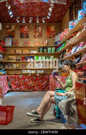 Chongqing, China - August 2019 : gelangweilte chinesische Verkäuferin, die sein Smartphone überprüft, während sie sitzt und auf Kunden in seinem Snack und Essen sh wartet Stockfoto