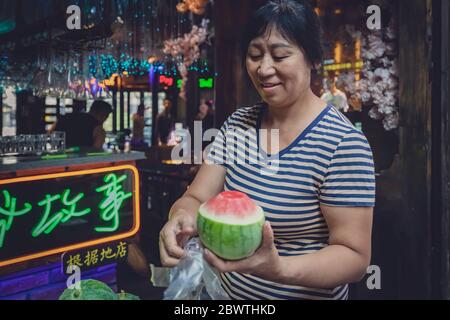 Feng Huang, China - August 2019 : Chinesische Frau mittleren Alters hält kleinen Schnitt offen köstliche und reife Wassermelone auf der Straße in der Altstadt in Feng Hu Stockfoto