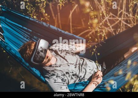 Junger Mann, der in der Hängematte entspannt und VR-Brillen verwendet Stockfoto