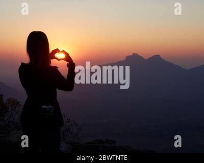 Silhouette einer Frau, die bei Sonnenuntergang mit ihren Händen das Herz formt, Serra da Leba, Angola Stockfoto