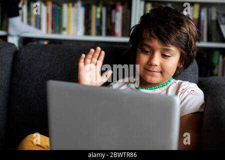 Niedlicher Junge winkt, während mit Laptop für Videoanruf im Wohnzimmer Stockfoto