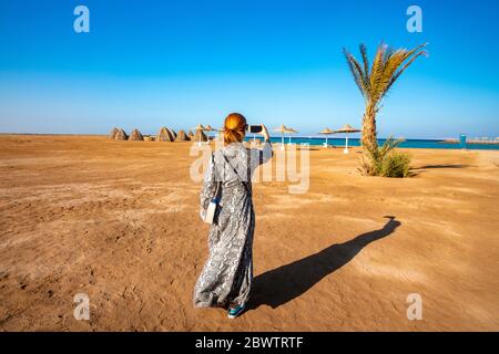 Ägypten, Hurghada, weibliche Touristen fotografieren Sonnenschirme und Strohhütten stehen entlang der Küste von Sahl Hasheesh Bucht Stockfoto