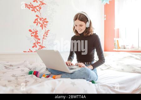 Portrait von Teenager-Mädchen mit Kopfhörer sitzen auf dem Bett mit Laptop Stockfoto