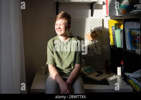 Portrait eines funkelnden Jungen, der zu Hause auf dem Schreibtisch sitzt und aus dem Fenster schaut