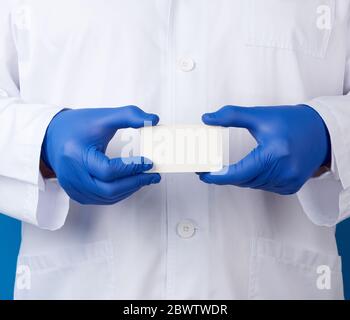 Arzt in einer weißen Robuniform und blauen sterilen Handschuhen gekleidet steht und hält leere weiße Papier Visitenkarte, Platz für die Inschrift Stockfoto