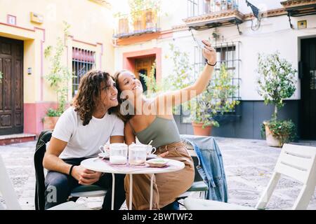 Junge Frau, die mit ihrem Freund Selfie über das Handy macht, während sie im Bürgersteig-Café, Santa Cruz, Sevilla, Spanien sitzt Stockfoto