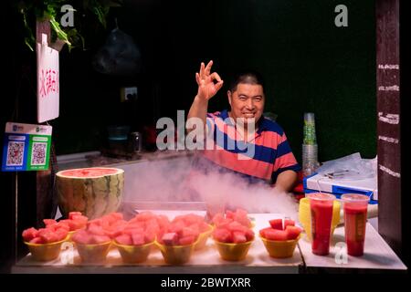 Chongqing, China - August 2019 : Glückliche chinesische Mann mittleren Alters Verkauf Portionen von frisch geschnitten saftige Wassermelone in CI Qi Kou Altstadt Stockfoto