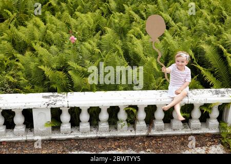 Kleines Mädchen auf einer Balustrade sitzend, das einen Ballon aus Pappe hält Stockfoto