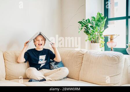 Portrait eines lachenden Jungen, der mit einem Buch auf der Couch im Wohnzimmer sitzt Stockfoto
