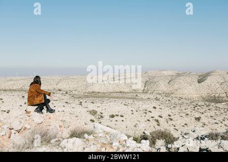 Die ganze Länge der Frau sitzt auf Felsen, während Blick auf Wüstenlandschaft gegen klaren blauen Himmel Stockfoto